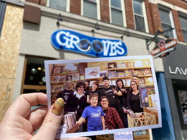 A printed photo of the staff and family of Grooves Record store is held up in front of the storefront