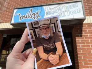 A printed photo of Milos Kral is held up in front of his storefront: Milos' Craft Beer Emporium