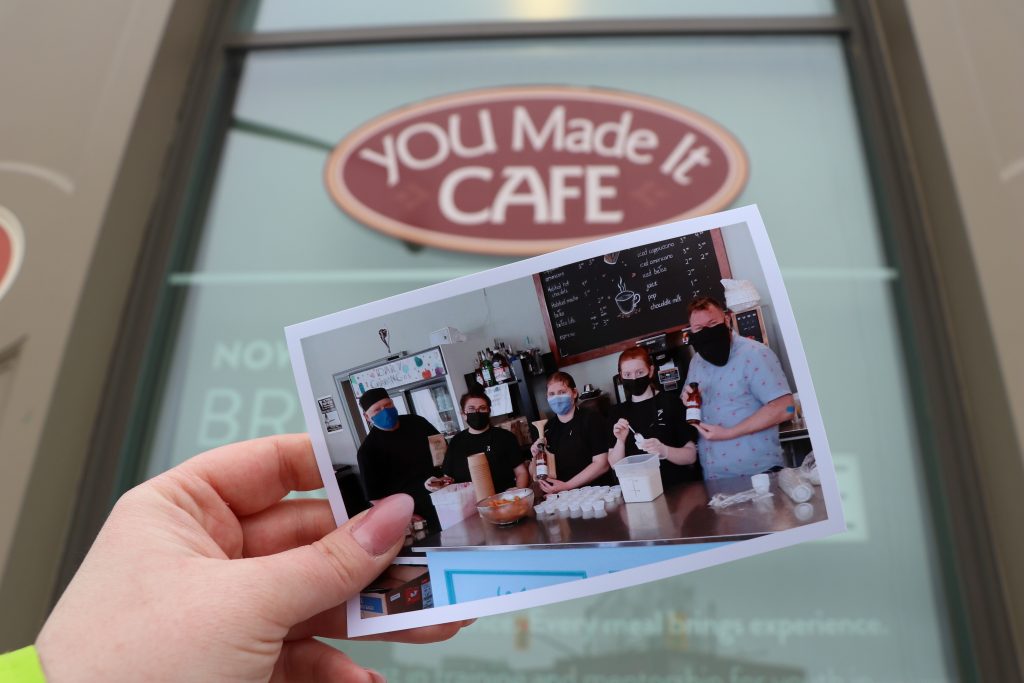 A printed photo of the staff is held in front of the You Made It Cafe.